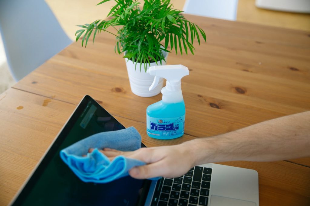 Close-up of hand cleaning a laptop screen with a blue cloth and spray bottle on wooden desk.
